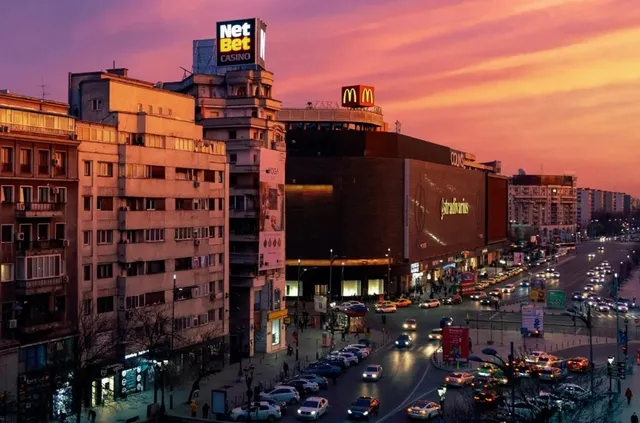 O peisaj urban la apus cu clădiri, trafic intens pe șoseauă și un cer în tonuri roz și portocaliu.