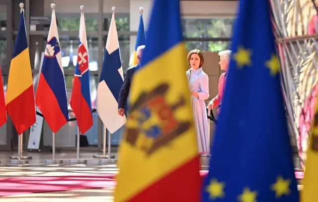 Două femei stau în picioare într-o încăpere cu mai multe steaguri colorate dispuse în rânduri.