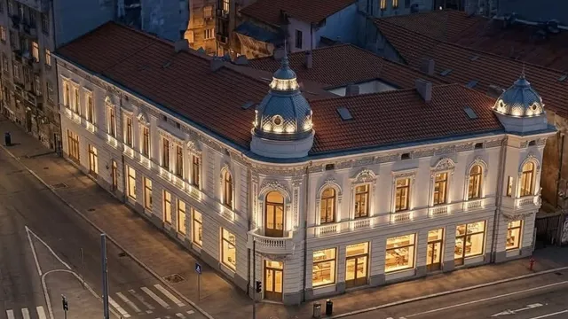 O clădire veche cu fațadă albă și acoperiș de țiglă roșie este iluminată de lumină caldă pe un strălucitor de seară.