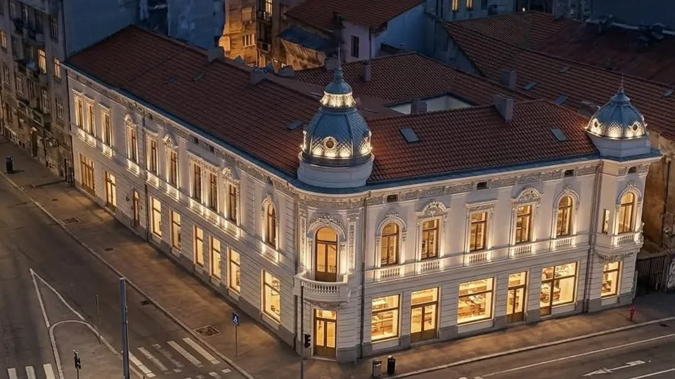 O clădire veche cu fațadă albă și acoperiș de țiglă roșie este iluminată de lumină caldă pe un strălucitor de seară.