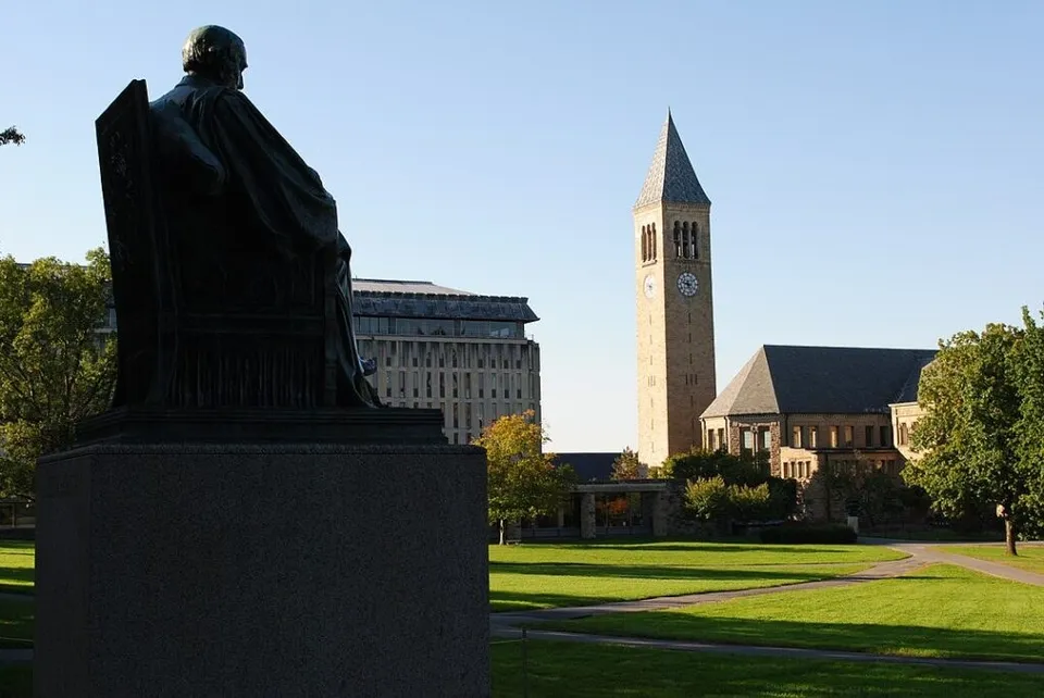 O statuie neagră de pe un soclu înalt este poziționată în prim-plan, având un turn cu ceas și clădiri universitare în fundal.
