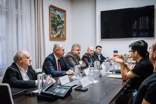 Cinci bărbați în costume și un bărbat în tricou negru stau la o masă lungă de conferință cu pahare de apă și telefoane pe ea.