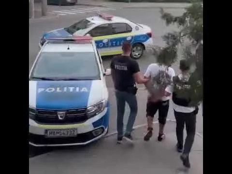 Doi polițiști însoțesc un bărbat încătușat, cu două mașini de poliție parcate în fundal, pe o stradă. 