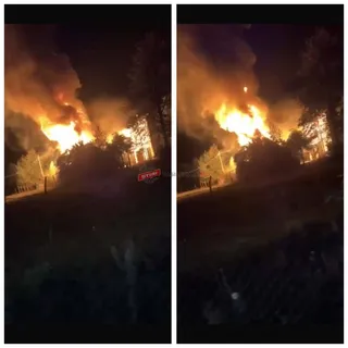 Flăcări mari și fum dens ies dintr-o structură de la noapte, înconjurate de vegetație și un gard de sârmă la prim-plan.