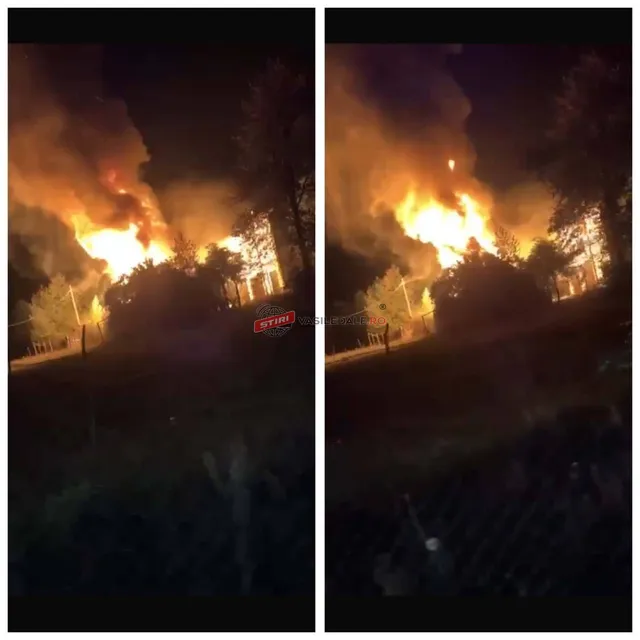 Flăcări mari și fum dens ies dintr-o structură de la noapte, înconjurate de vegetație și un gard de sârmă la prim-plan.