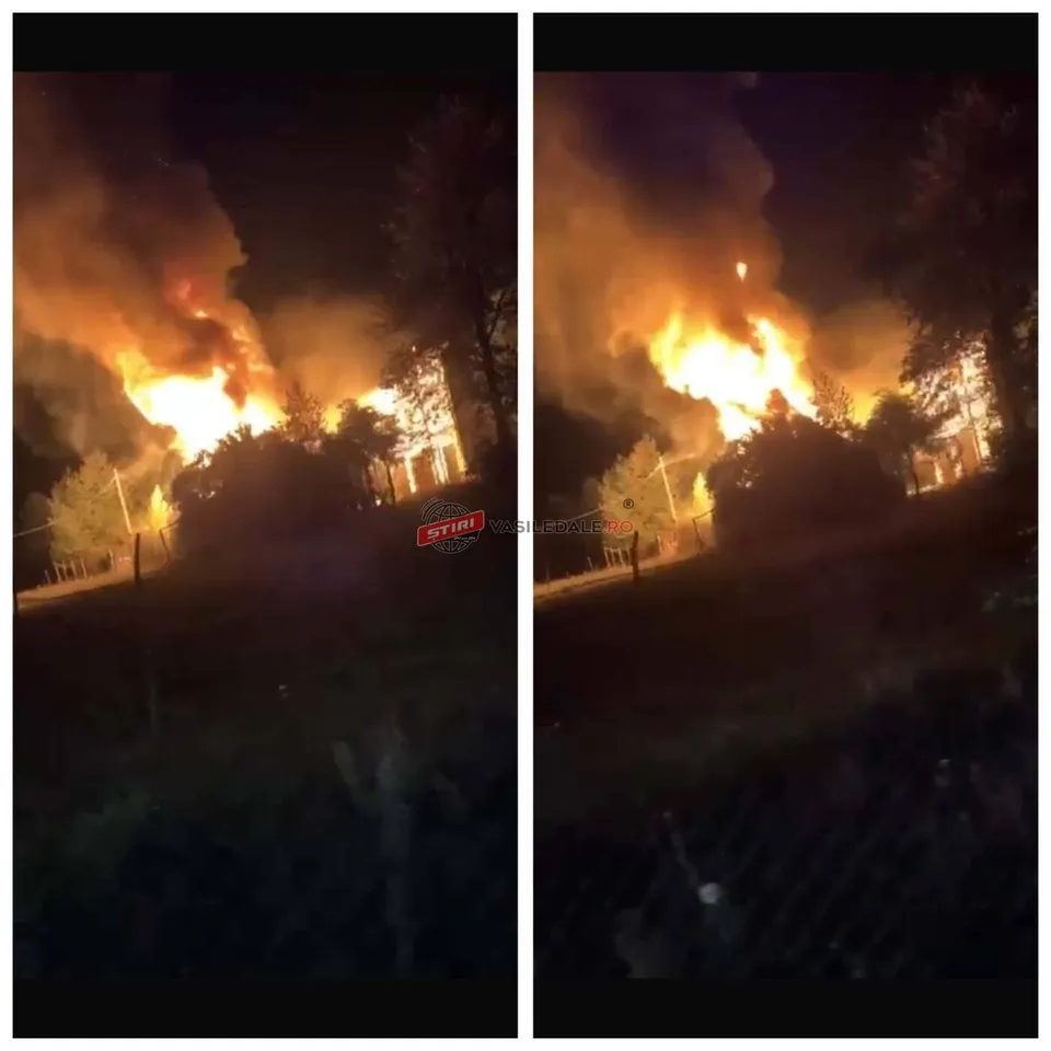 Flăcări mari și fum dens ies dintr-o structură de la noapte, înconjurate de vegetație și un gard de sârmă la prim-plan.