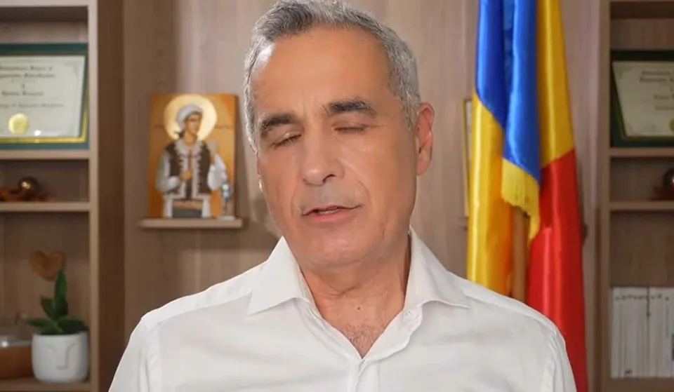 Un bărbat cu părul cărunt și cămașă albă stă în fața unui raft cu diplome, o icoană și un steag colorat.