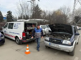 Un bărbat în vestă albastră și blugi stă între un van alb cu portbagajul deschis și o mașină neagră cu capota ridicată.