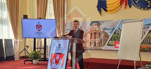 Un bărbat într-un costum închis la culoare stă în spatele unui podium lângă un ecran și un panou cu o imagine a unei clădiri.