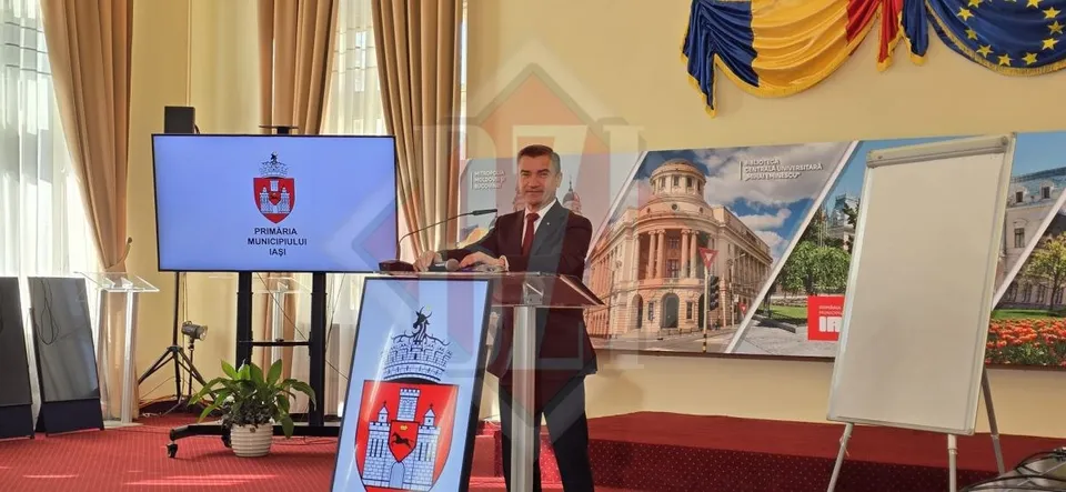 Un bărbat într-un costum închis la culoare stă în spatele unui podium lângă un ecran și un panou cu o imagine a unei clădiri.