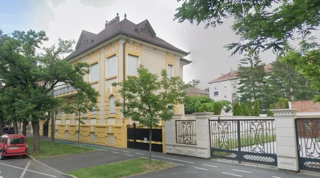 O clădire galbenă cu acoperiș închis este situată lângă un gard alb cu porți metalice negre și copaci verzi.