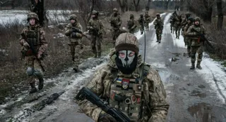 Soldați în uniforme de camuflaj și echipament militar merg pe un drum de țară înconjurat de zăpadă și copaci lipsiți de frunze.