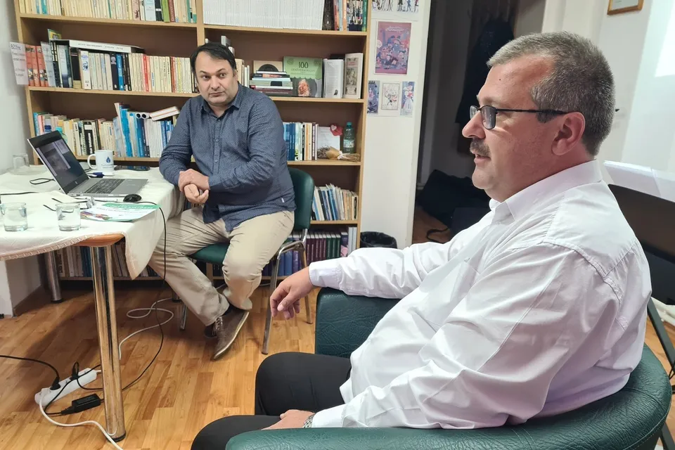 Un bărbat cu ochelari și cămașă albă stă pe un scaun verde, în timp ce un alt bărbat stă la o masă lângă un laptop.