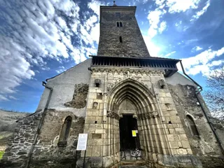 O clădire veche din piatră prezintă un portal gotic cu arcade multiple și un turn înalt sub un cer cu nori.
