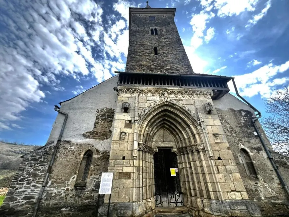 O clădire veche din piatră prezintă un portal gotic cu arcade multiple și un turn înalt sub un cer cu nori.