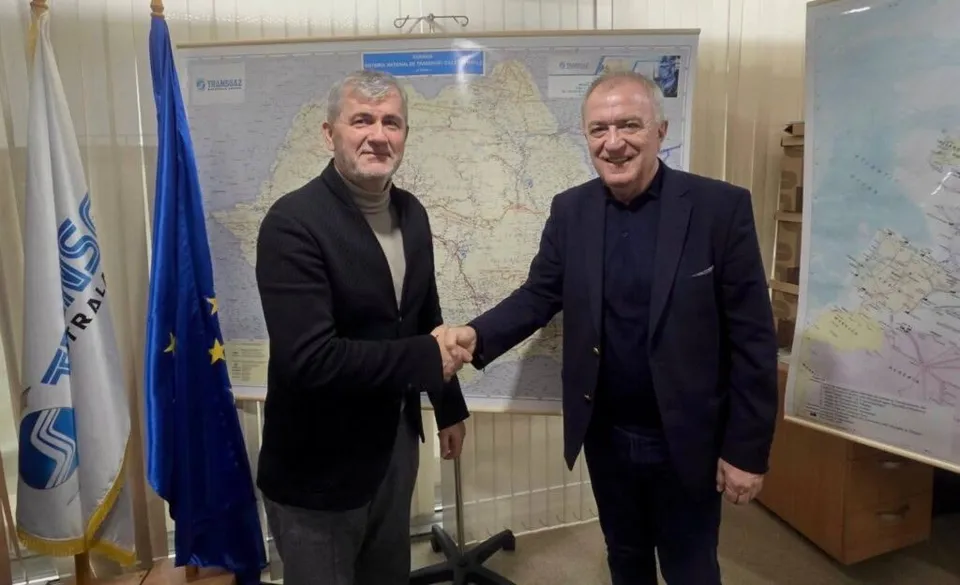 Doi bărbați stau în picioare în fața unei hărți și își strâng mâinile, unul purtând sacou negru și celălalt sacou albastru închis.