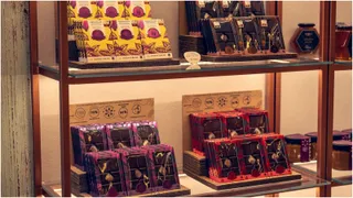 Diverse cutii de ciocolată și borcane cu produse alimentare sunt expuse pe rafturi de sticlă într-un vitrină de lemn.
