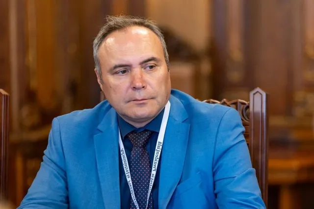 Un bărbat cu părul scurt și costum albastru poartă o cămașă închisă la culoare, o cravată și un cordon alb în jurul gâtului.