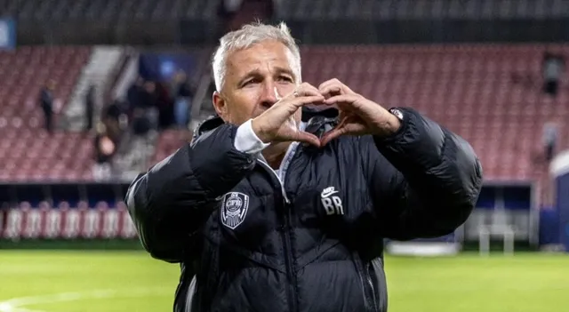Un bărbat cu părul cărunt, purtând o jachetă neagră cu un logo, face semnul unei inimi cu mâinile pe un teren de fotbal.
