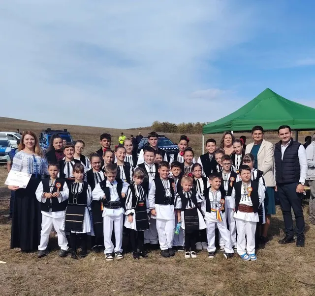 Un grup de copii și adulți în costume tradiționale albe și negre stau în rând pe un câmp sub un cer senin.