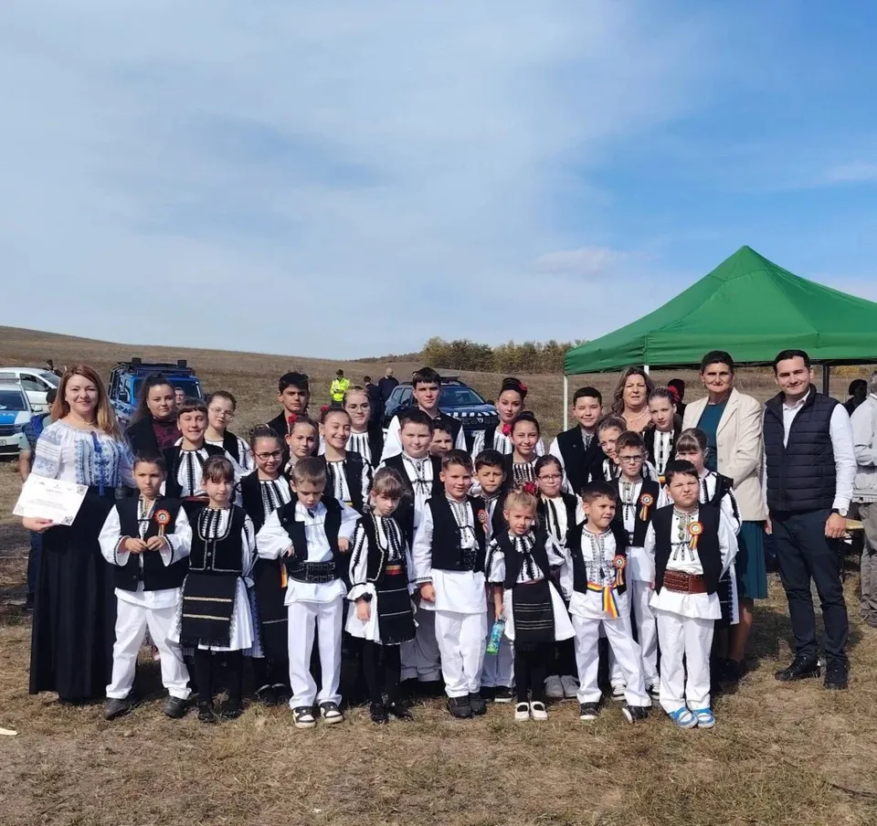 Un grup de copii și adulți în costume tradiționale albe și negre stau în rând pe un câmp sub un cer senin.