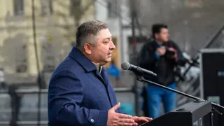Un bărbat cu părul scurt și haine închise la culoare stă în fața unui microfon de pe un podium.