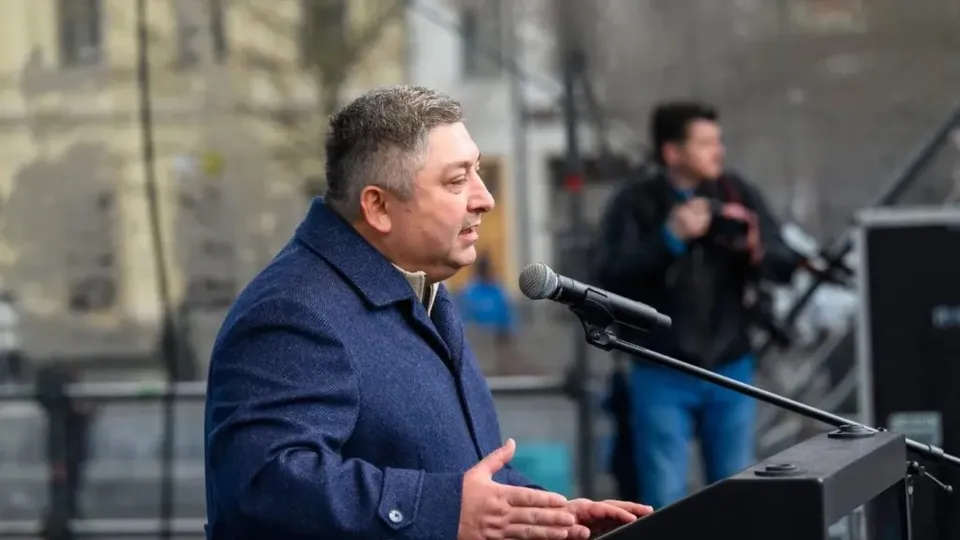 Un bărbat cu părul scurt și haine închise la culoare stă în fața unui microfon de pe un podium.