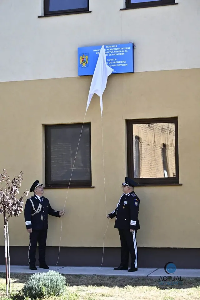 Doi bărbați în uniforme închise ridică un steag alb folosind două sfori în fața unei clădiri cu ferestre și un panou albastru.