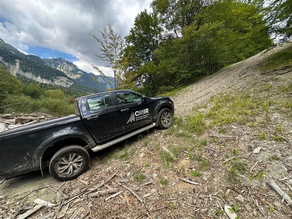 Un pickup negru cu inscripții albare este parcat pe un teren accidentat, înconjurat de vegetație și copaci, sub un cer înnorat.