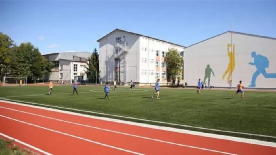 Câțiva copii joacă pe un teren de fotbal cu iarbă artificială, lângă o pistă de atletism roșie și clădiri de școală.