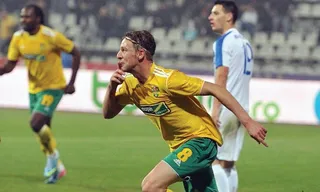 Un jucător de fotbal în echipament galben și verde aleargă pe un teren de gazon verde, în timp ce alți doi jucători sunt vizibili în fundal.