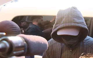 O persoană cu capota unui hanorac gri și o șapcă albă acoperă fața, în timp ce un grup de oameni este vizibil într-un vehicul în fundal.