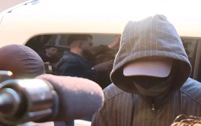 O persoană cu capota unui hanorac gri și o șapcă albă acoperă fața, în timp ce un grup de oameni este vizibil într-un vehicul în fundal.