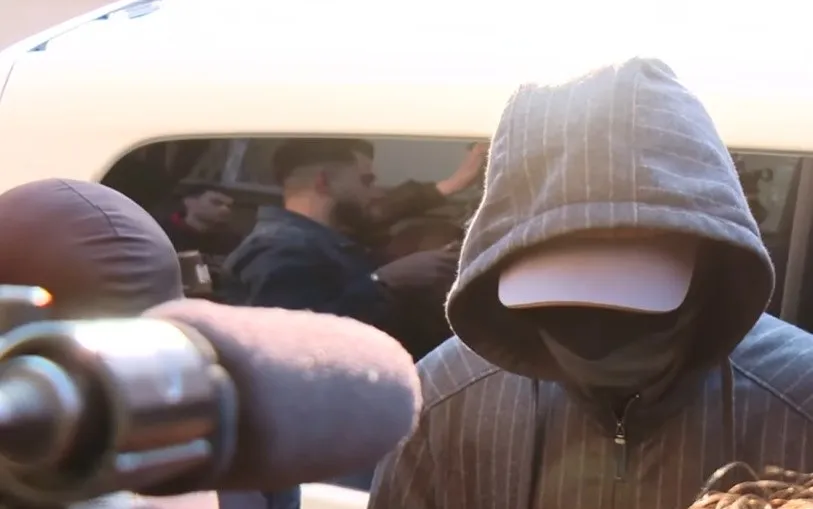 O persoană cu capota unui hanorac gri și o șapcă albă acoperă fața, în timp ce un grup de oameni este vizibil într-un vehicul în fundal.