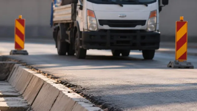 Un camion alb circulă pe un drum asfaltat între două conuri de semnalizare galben-roșii, lângă o bordură de beton.
