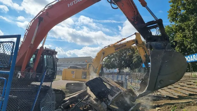 O excavatoare portocalie de mărime mare folosește un cupă pentru a manipula resturi de materiale pe un șantier sub un cer cu nori.