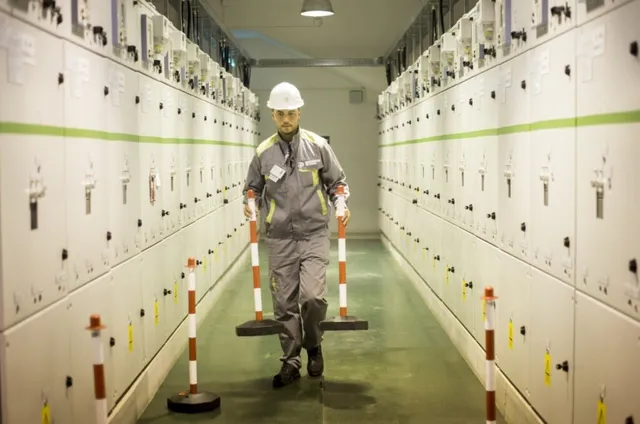 Un bărbat cu cască albă și echipament de lucru gri merge printr-un coridor dintre panouri electrice albe de înaltă tensiune.