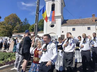 Oameni îmbrăcați în costume populare dansează pe un drum în fața unei clădiri albe cu un turn și două steaguri colorate.