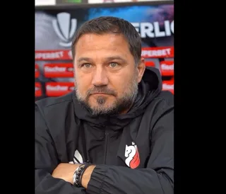 Un bărbat cu barbă și păr scurt, purtând o jachetă neagră cu un logo alb și roșu, stă cu brațele încrucișate în fața unui fundal colorat.