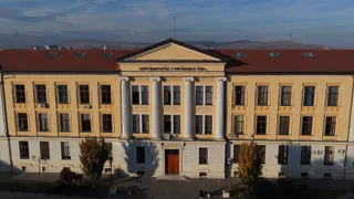 O clădire galbenă cu trei etaje prezintă un corp central cu coloane albe și un acoperiș roșu sub un cer senin.