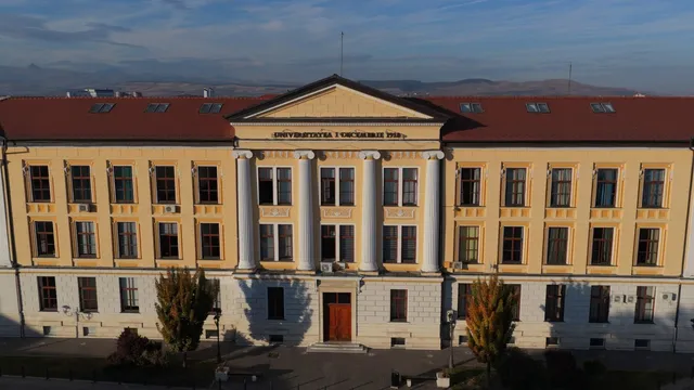 O clădire galbenă cu trei etaje prezintă un corp central cu coloane albe și un acoperiș roșu sub un cer senin.