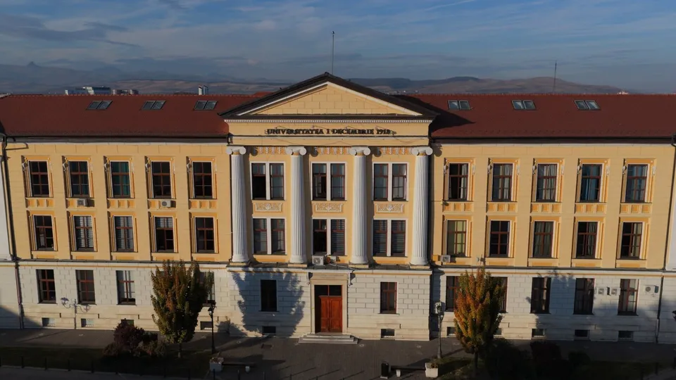 O clădire galbenă cu trei etaje prezintă un corp central cu coloane albe și un acoperiș roșu sub un cer senin.