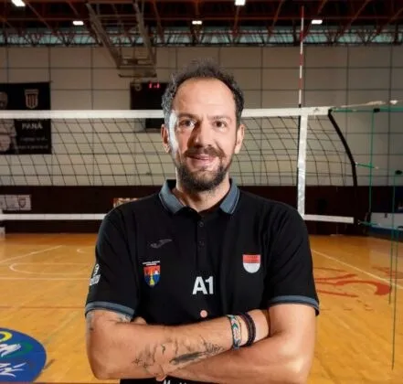 Un bărbat cu barbă și păr scurt stă cu brațele încrucișate într-o sală de sport cu un teren de volei în fundal.