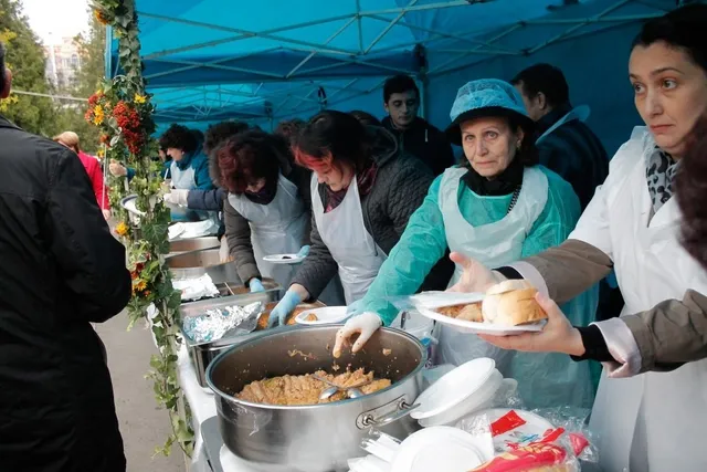 Femei cu șorțuri și bonete distribuie mâncare din oale mari pe farfurii de plastic unor persoane aflate la o masă sub un cort albastru.