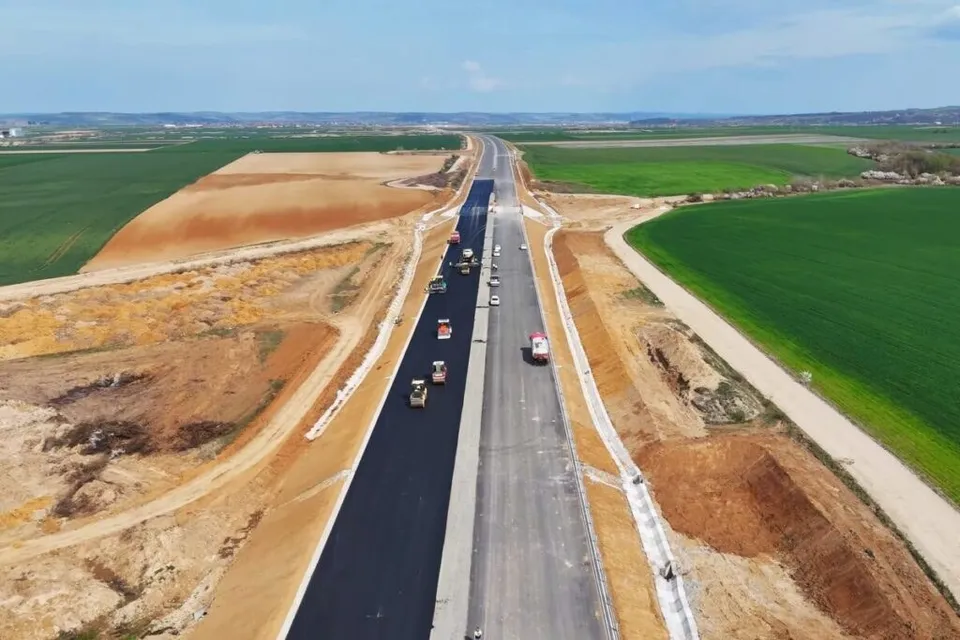 O vedere aeriană prezintă două caraje de autostradă paralele, unul cu asfalt negru și unul gri, înconjurate de terenuri agricole și zone de excavare.