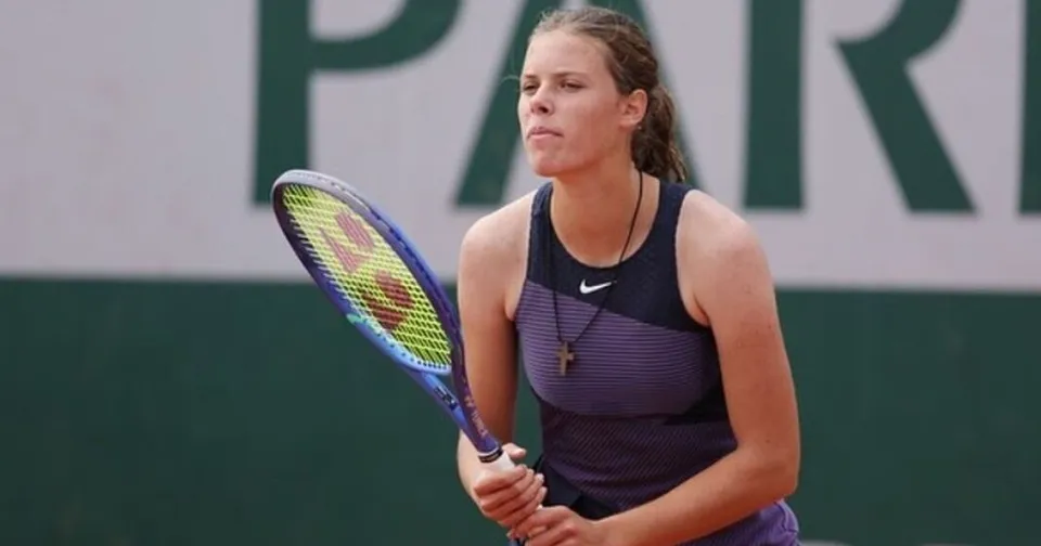 O tânără sportivă cu părul împletit poartă o rochie sportivă violet și ține o rachetă de tenis în mâini pe un teren de argilă.