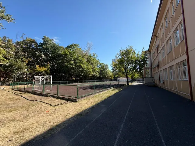 Un teren de sport cu gard de sârmă și un por de fotbal se află lângă o clădire înaltă și o pistă asfaltată.