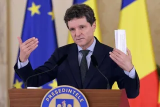 Un bărbat într-un costum închis la culoare și cravată stă în spatele unui podium, ridicând mâna stângă cu o foaie de hârtie albă.