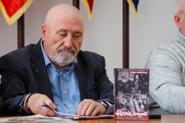 Un bărbat cu barbă căpătată și sacou închis se apleacă peste o masă, scriind pe o foaie de hârtie lângă o carte cu copertă neagră.