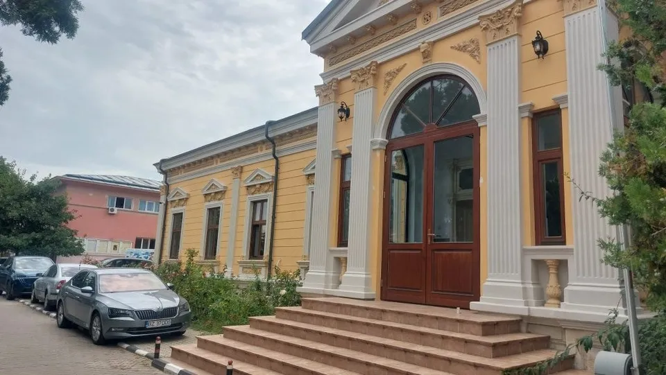 O clădire galbenă cu detalii albe, uși duble de lemn și scări de piatră este vizibilă sub un cer înnorat.